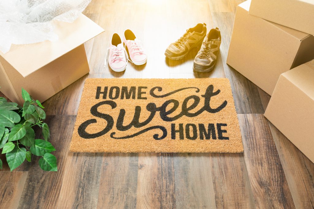 Home Sweet Home Welcome Mat, Moving Boxes, Women and Male Shoes and Plant on Hard Wood Floors.